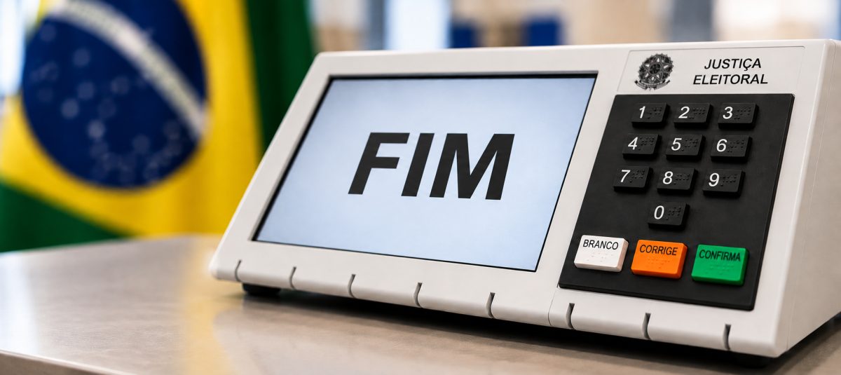 Urna eletrônica da Justiça Eleitoral sobre uma mesa, com a palavra FIM na tela e a bandeira do Brasil desfocada ao fundo